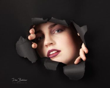 portret van jonge vrouw kijkend door gescheurd zwart papier een gedeelte van haar gezicht is zichtbaar terwijl haar vingers het papier vasthouden